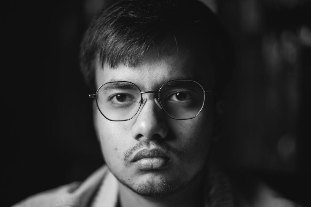 Close-up portrait of a young man wearing glasses, staring ahead with a blank expression, representing emotional disconnection and difficulty identifying or expressing feelings.
