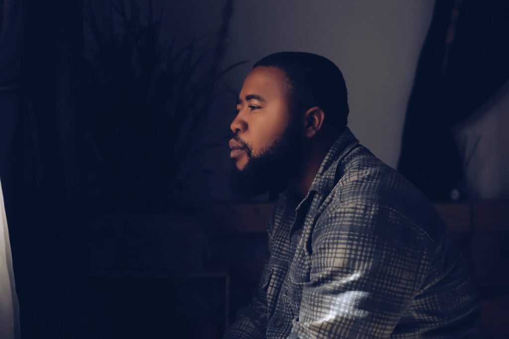 Man sitting alone in low light, gazing toward a window with a thoughtful, distant expression, symbolizing the emotional struggle and internal disconnect often experienced by men with alexithymia.