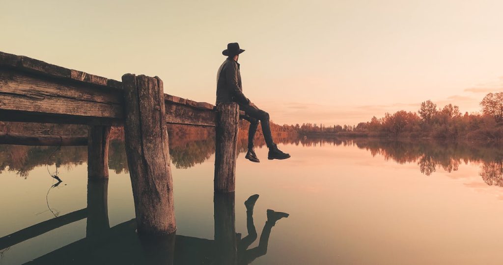 Man sitting alone on an old wooden pier at sunset, reflecting in silence over a calm lake.