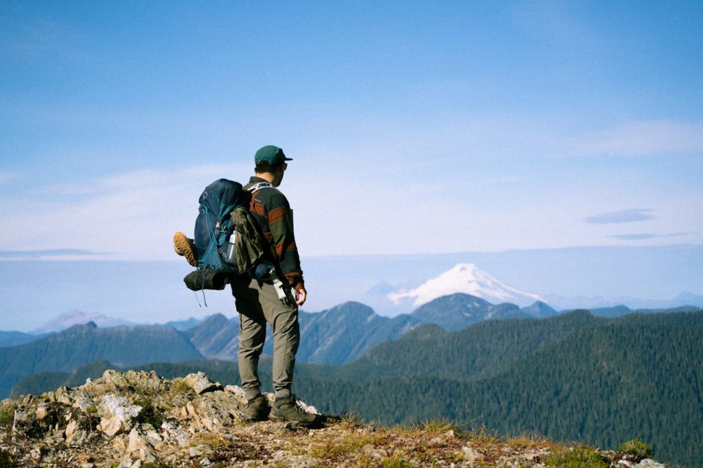 Man standing on a mountain ridge at sunrise with a large hiking backpack, looking out over distant peaks and a snow-covered summit, symbolizing solitude, clarity, and personal growth.