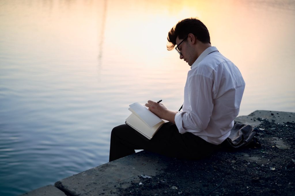 Man journaling alone by the water at sunset, reflecting in silence and writing in a notebook.