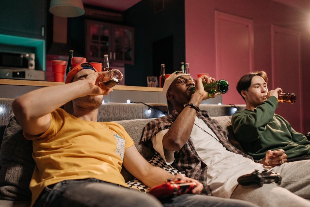 Three young men drinking beer together on a couch, engaging in casual behavior that masks deeper emotional disconnection.