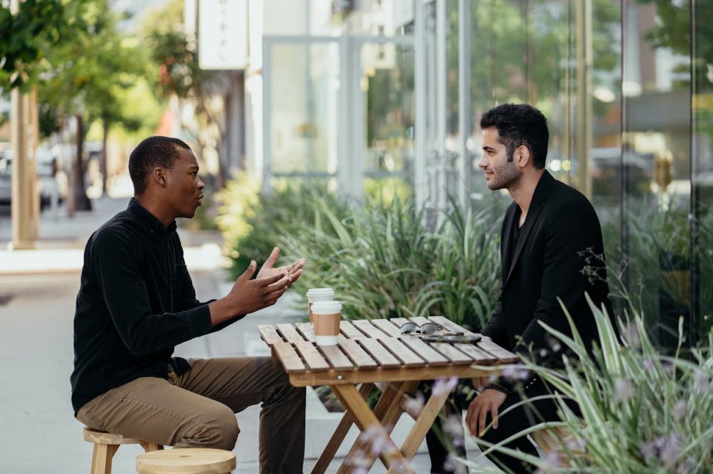 Two men talking openly at an outdoor café, illustrating supportive conversations that encourage men to seek therapy.