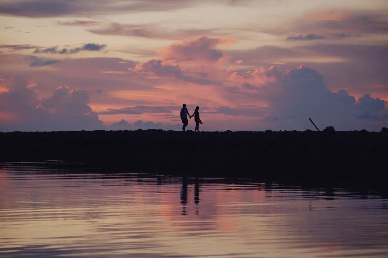 Couple holding hands while walking along a shoreline at sunset, silhouetted against a sky of soft pink and purple clouds, symbolizing shared curiosity and emotional connection.