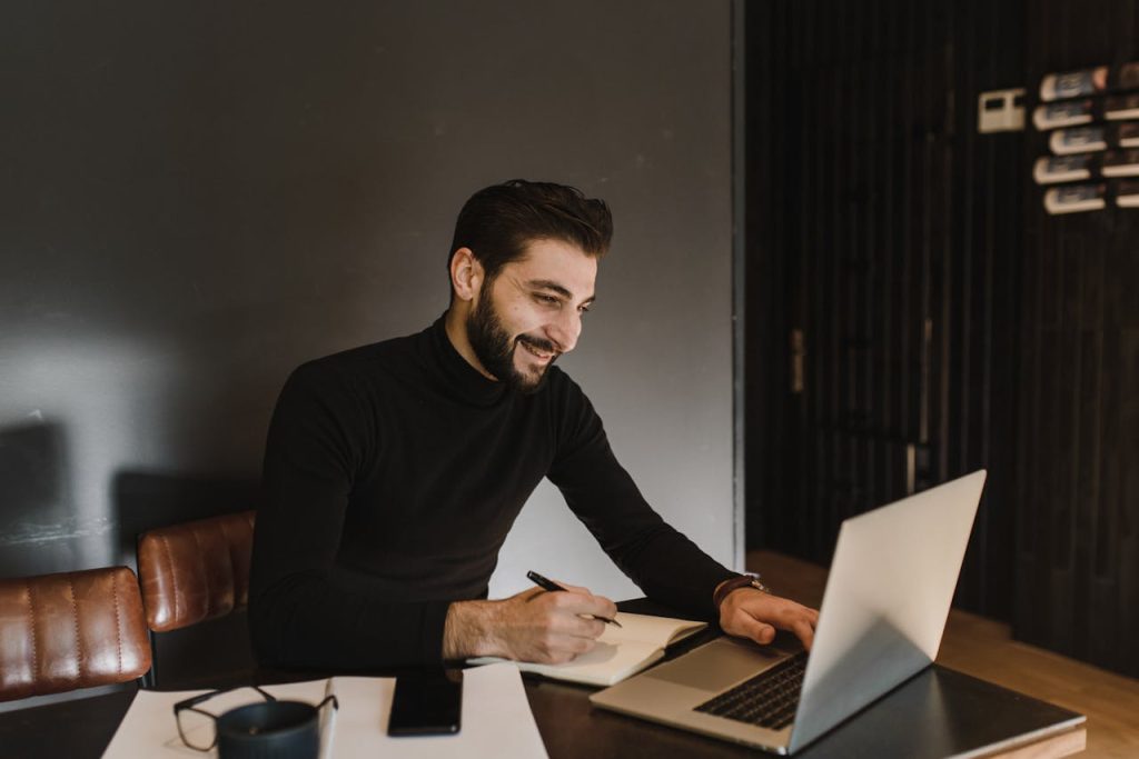 Man smiling while working on a laptop and writing in a notebook, displaying focused and mindful productivity.