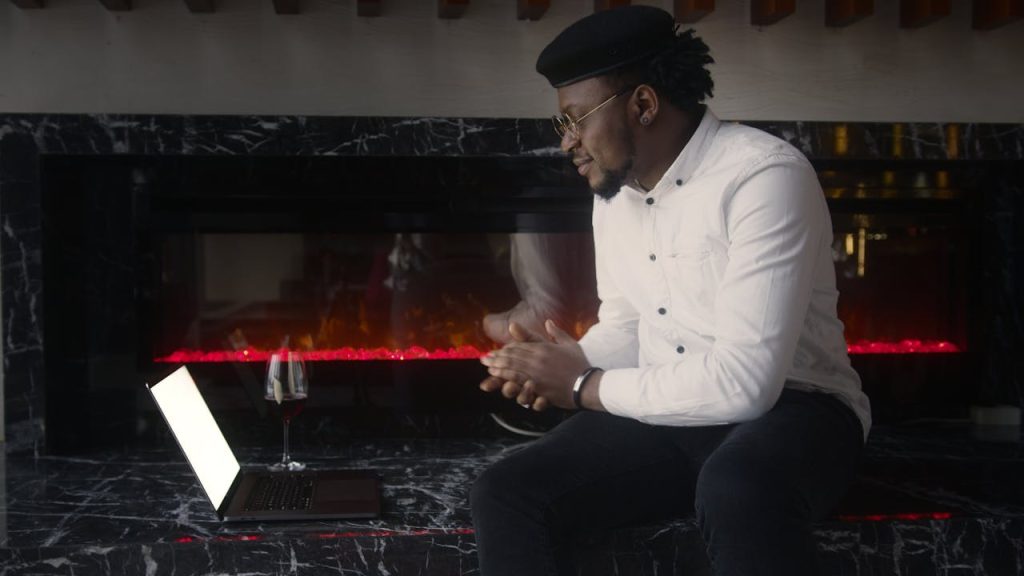 A man sitting by a fireplace engaged in a video call on his laptop, symbolizing the importance of male support groups through accessible online connection.