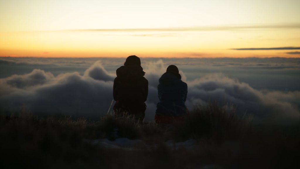 Two people sitting together in silence above the clouds during sunset, peacefully unplugged and sharing a quiet moment.