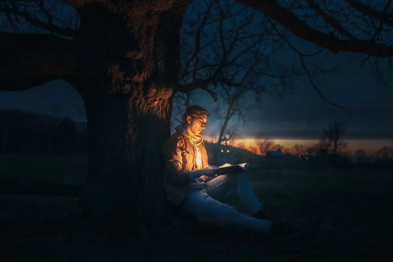 Man reading under a tree at dusk with warm light illuminating his face, symbolizing introspection, self-reflection, and the inner work essential to authentic masculinity.