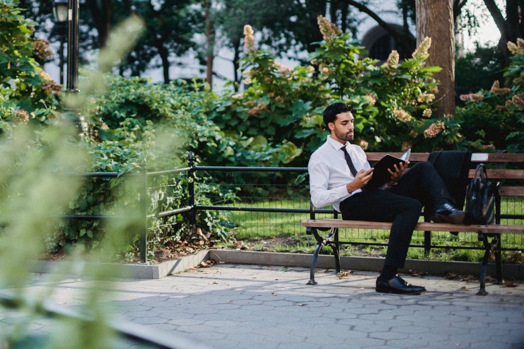 Man in business attire sitting on a park bench reading a notebook, taking a break from work to create healthy boundaries between professional and personal life.
