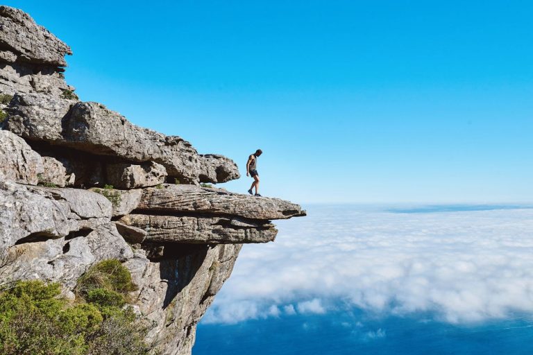 Man standing on the edge of a mountain cliff above the clouds, symbolizing the balance between ambition and inner peace.