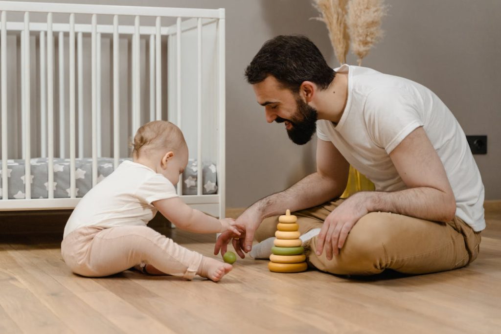 Father sitting on the floor playing with his baby, guiding a stacking toy during the first year of parenthood and bonding through early development.