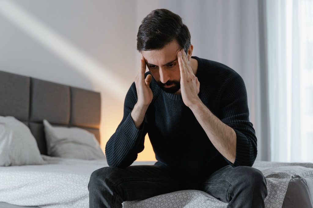 A man sitting on the edge of his bed with his head in his hands, appearing overwhelmed—illustrating the need for emotional “first aid” for men during tough times.