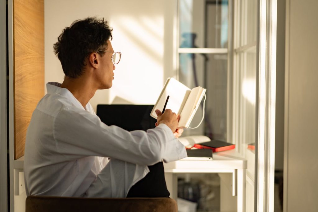 A man sitting by a window journaling in a notebook, reflecting quietly as sunlight fills the room—representing emotional self-awareness and healthy introspection.