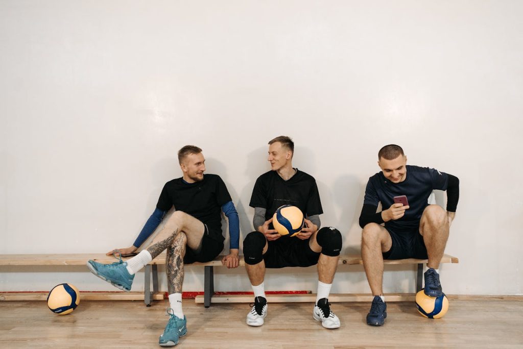 Three men sitting on a gym bench with volleyballs, talking and laughing after practice, showing healthy masculine camaraderie.