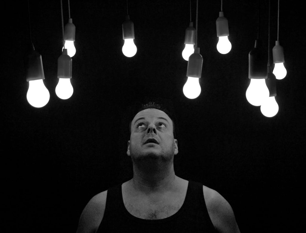 Man looking up at glowing lightbulbs in a dark room, symbolizing an aha moment sparked by boredom and mental stillness.