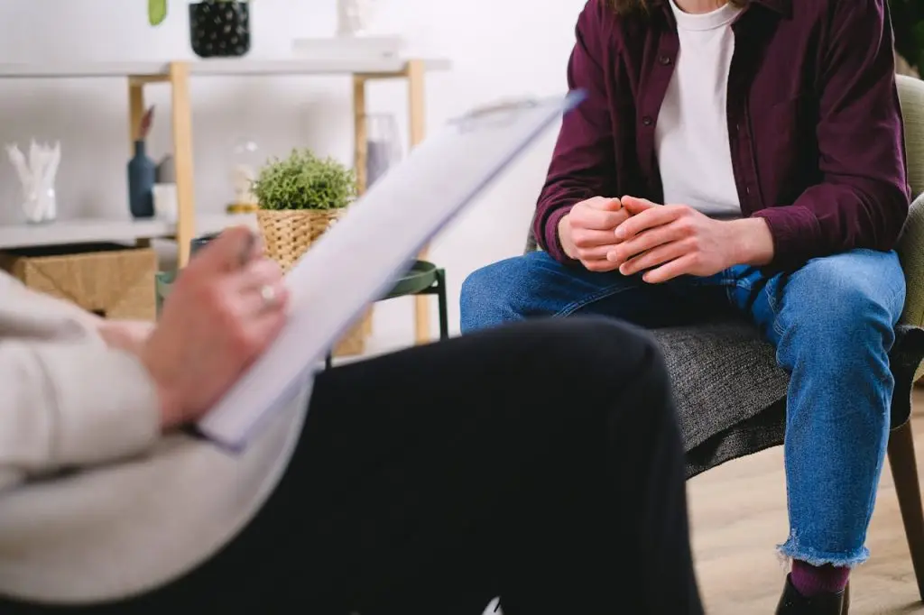 Man sitting in a therapy session, hands clasped while speaking with a counselor taking notes.