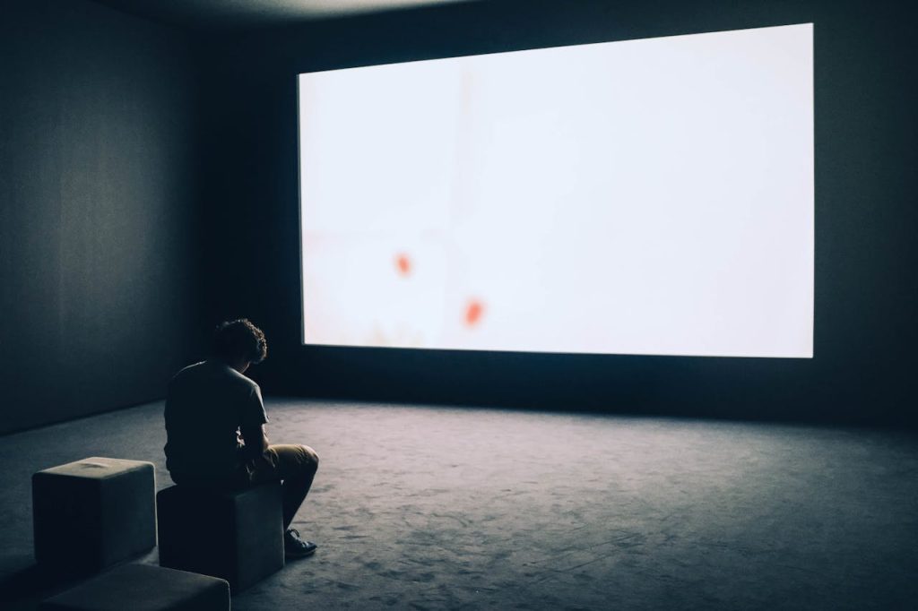 A man sitting alone in a dark room facing a bright screen, symbolizing isolation and compulsive behavior linked to porn addiction.