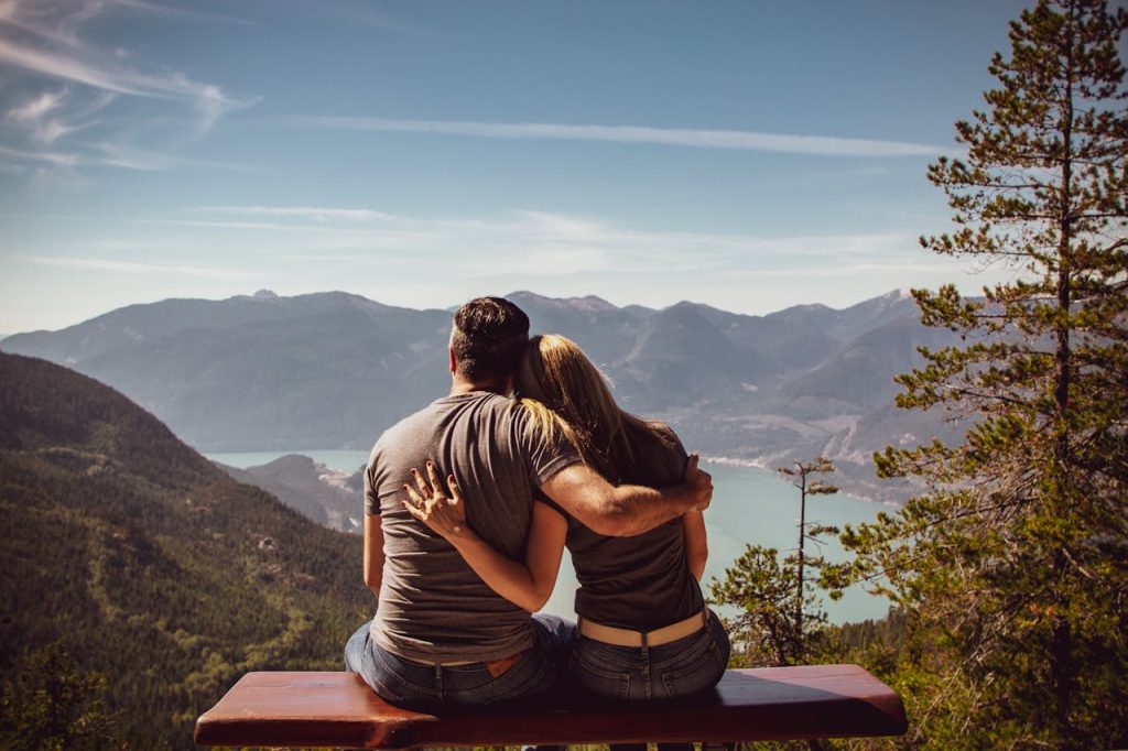 A couple sitting closely on a bench overlooking a mountain landscape, the man embracing his partner, symbolizing emotional attunement and understanding her emotions in relationships.