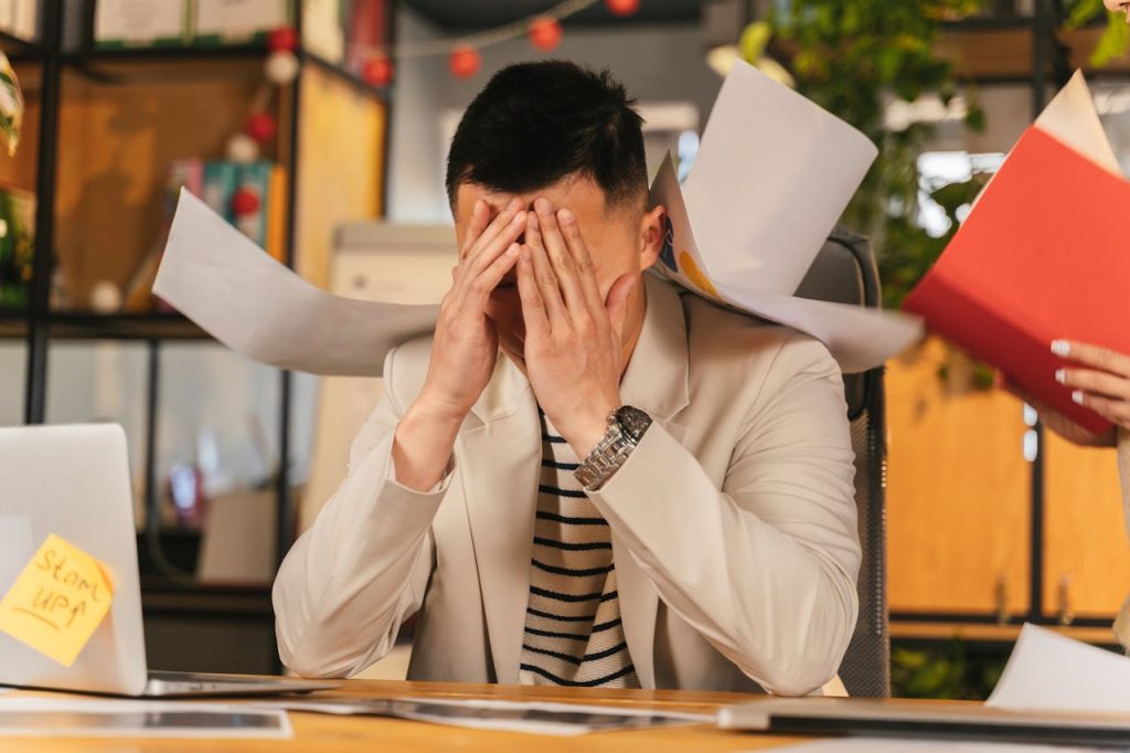 Overwhelmed man at his desk holding his head in his hands while papers fly around him, symbolizing toxic self-reliance and the struggle of trying to carry everything alone.