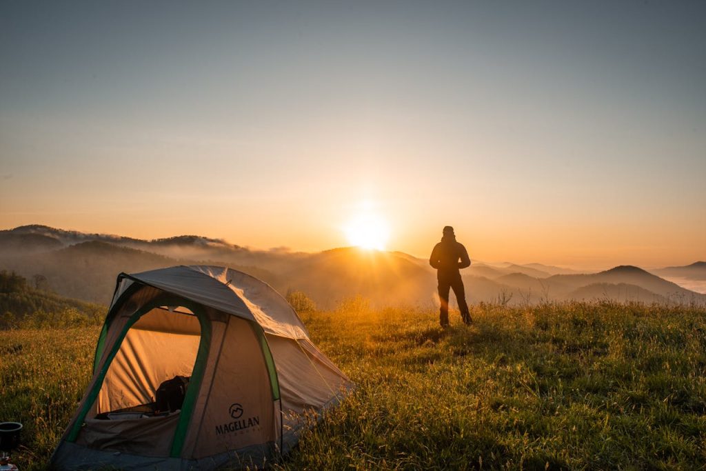 Man standing beside a tent at sunrise, looking out over peaceful mountain scenery.