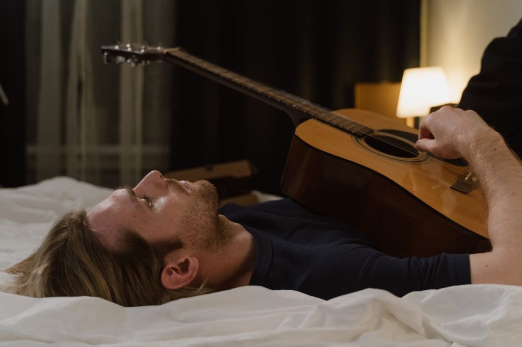 A man lying on a bed playing an acoustic guitar, representing creative outlets for men through music, solitude, and emotional expression.