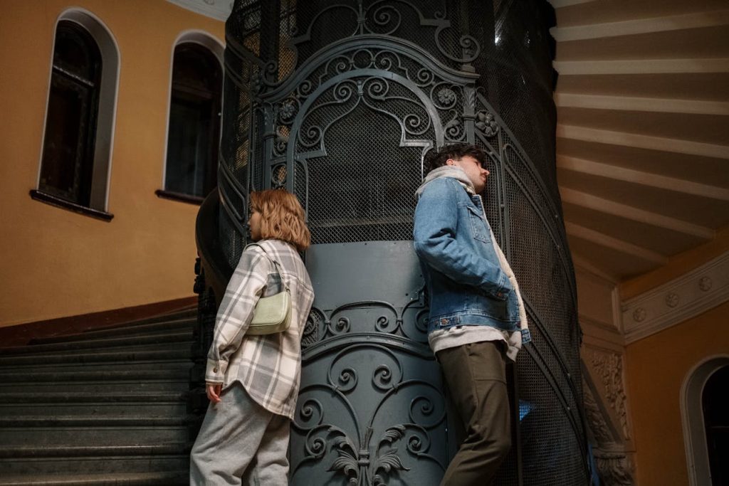 Couple standing back-to-back in a vintage stairwell, avoiding eye contact, symbolizing emotional disconnection and unresolved attachment patterns in adult relationships.