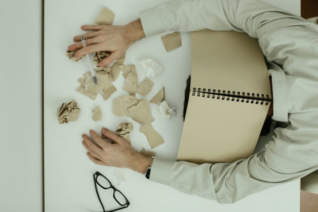 A man overwhelmed at his desk with his head down and papers scattered around him, symbolizing the exhaustion caused by chronic people pleasing.