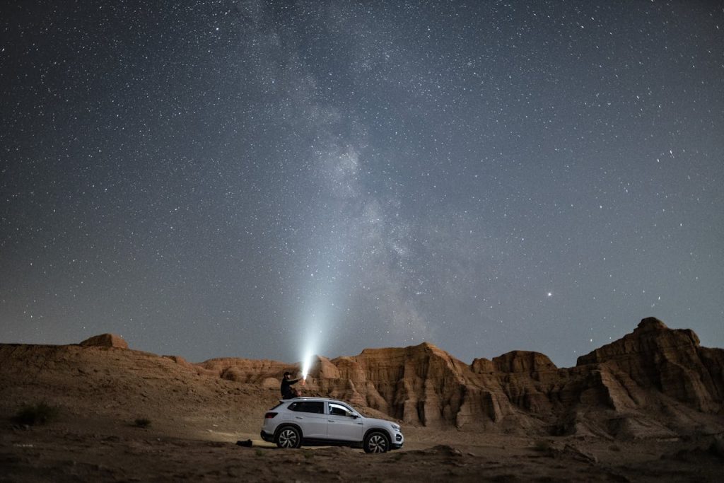 A man standing on a car in a desert at night, shining a light toward the Milky Way, representing creative outlets for men through exploration and photography.