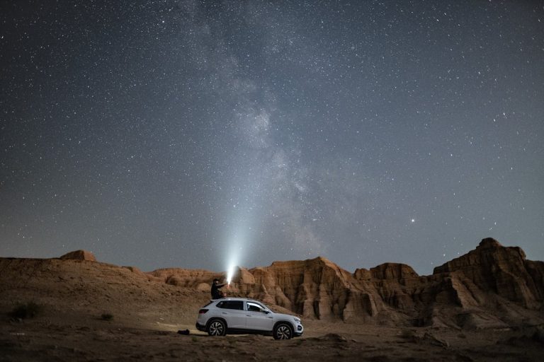 A man standing on a car in a desert at night, shining a light toward the Milky Way, representing creative outlets for men through exploration and photography.