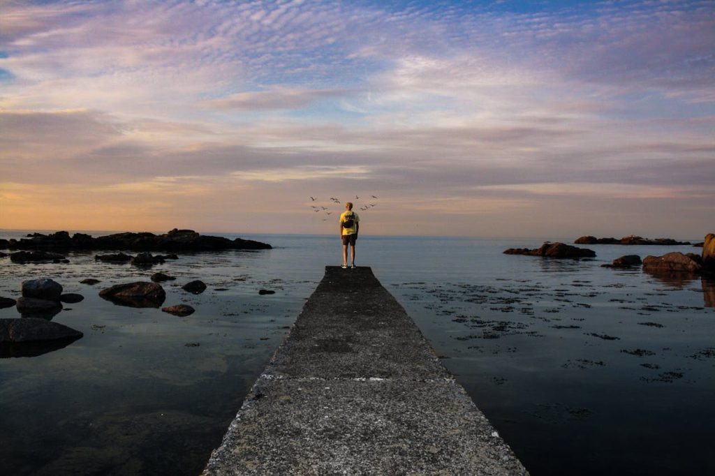 Person standing alone at the end of a pier overlooking a calm sea at sunset, symbolizing the difference between solitude and loneliness.