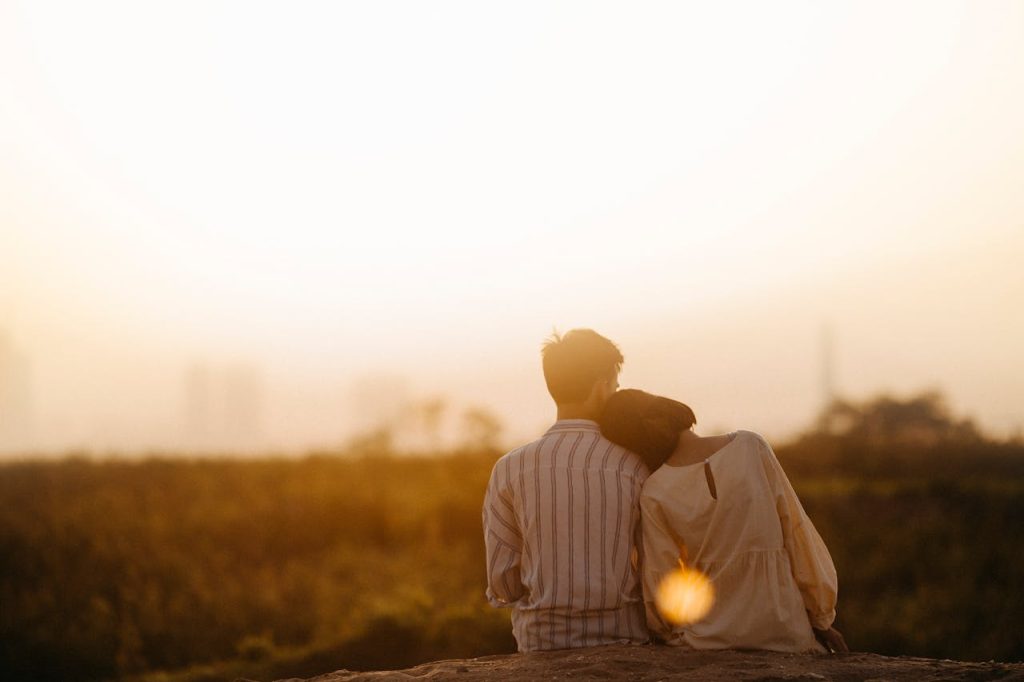A couple sitting close together at sunset, the woman resting her head on the man’s shoulder, symbolizing emotional presence, listening, and understanding her emotions.