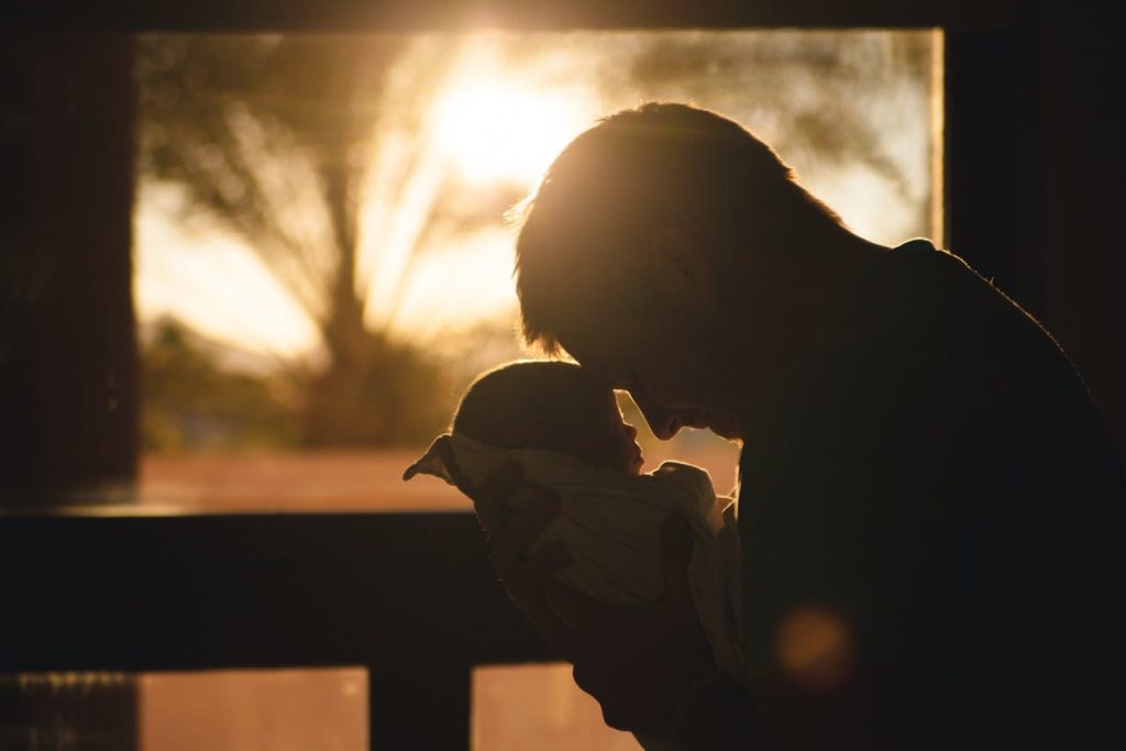 Father gently holding newborn at sunrise, showing emotional strength and tenderness while redefining manly strength.