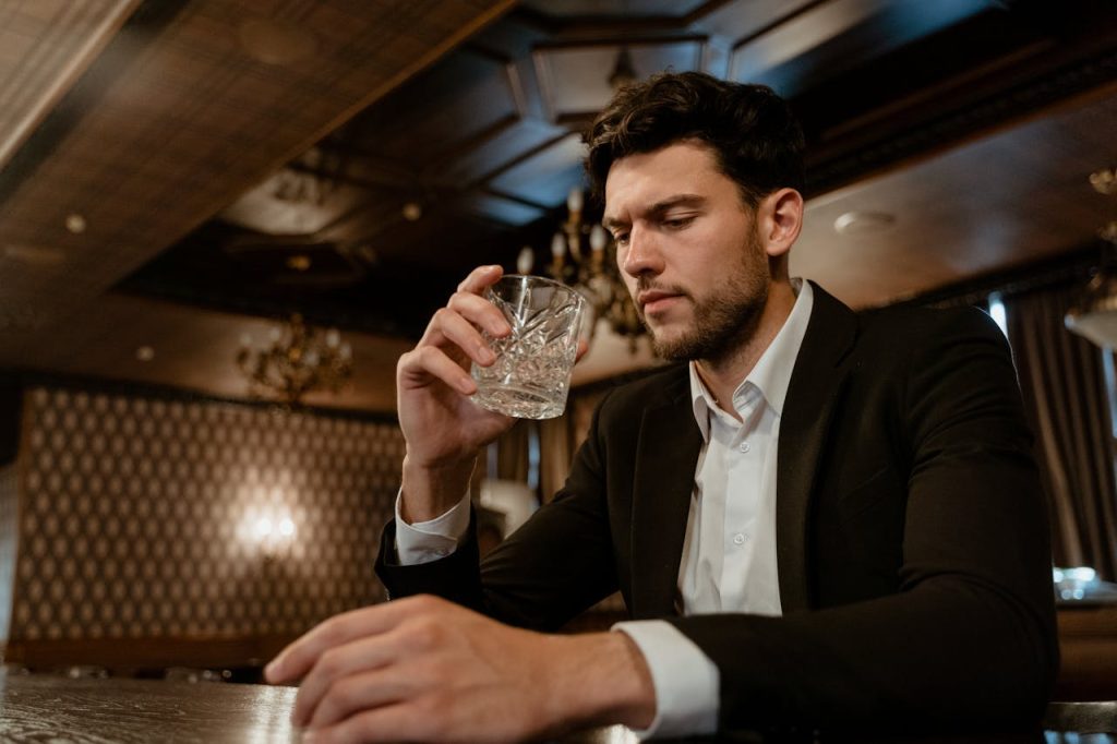 Man sitting alone at a table holding a glass, appearing contemplative, symbolizing how childhood trauma can influence men’s mental health and coping behaviors.