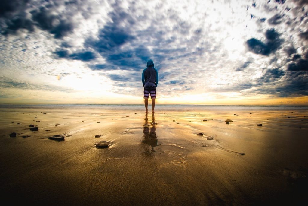 Person standing alone on a beach at sunrise, calmly observing the sky like watching emotions pass by.