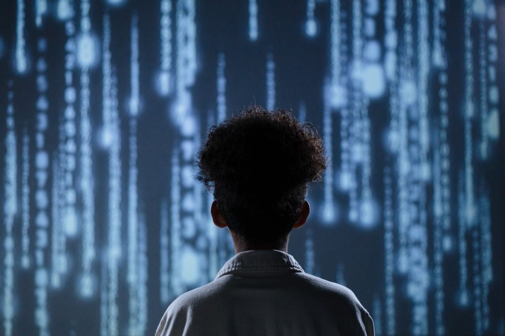 Young boy standing before a digital wall of code, symbolizing early programming that teaches boys to suppress vulnerability and avoid asking for help.