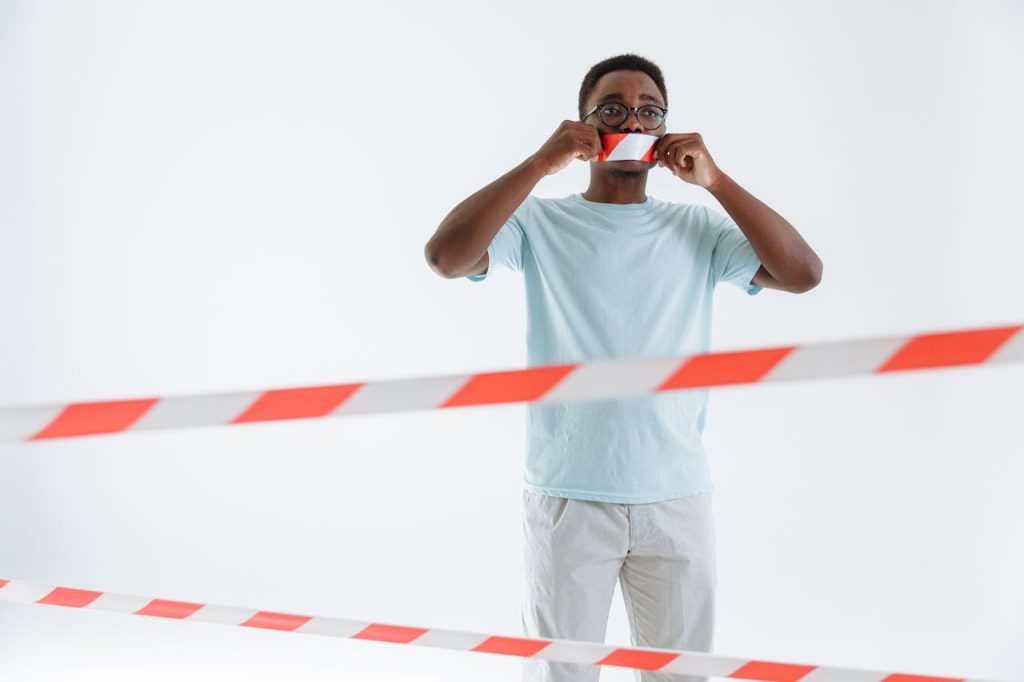 Man placing tape over his mouth while standing behind caution lines, symbolizing men’s mental health stigma and the pressure to stay silent.