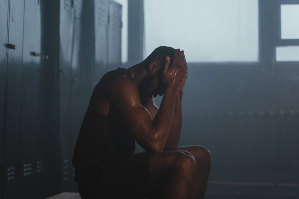 Man sitting exhausted in a locker room with his head in his hands, illustrating redefining manly strength beyond physical toughness.