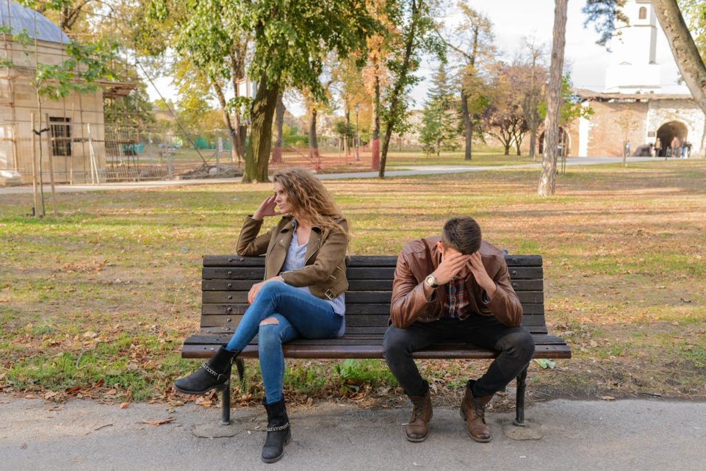 A couple sitting apart on a park bench, the man holding his head in distress while the woman looks away, illustrating emotional misunderstanding and difficulty understanding her emotions.