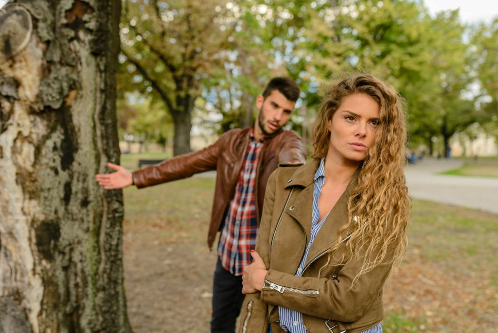 Couple arguing in a park, woman looking emotionally distant while the man reaches toward her, symbolizing how unresolved childhood patterns influence adult romantic relationships.