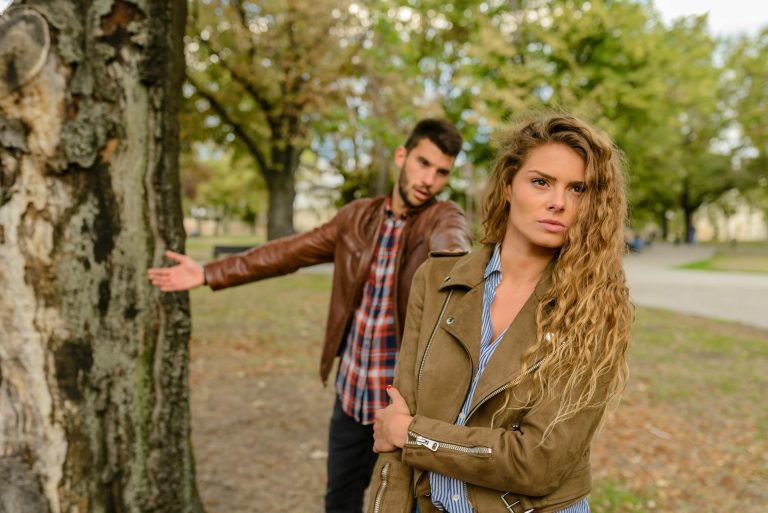 Couple arguing in a park, woman looking emotionally distant while the man reaches toward her, symbolizing how unresolved childhood patterns influence adult romantic relationships.