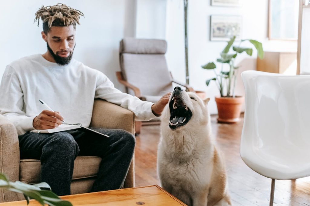 A man journaling in a calm living room while gently petting his dog, symbolizing reflection, emotional healing, and self-forgiveness.