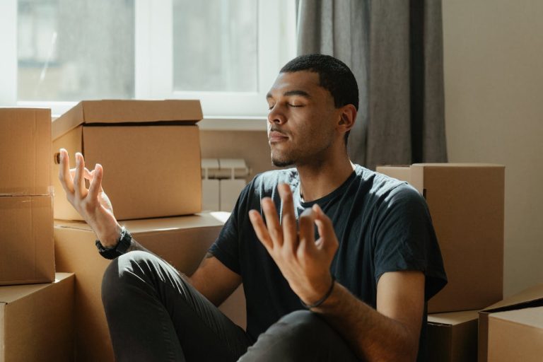 A man sitting calmly among moving boxes with eyes closed and hands open, practicing breathing for anger management and emotional regulation.