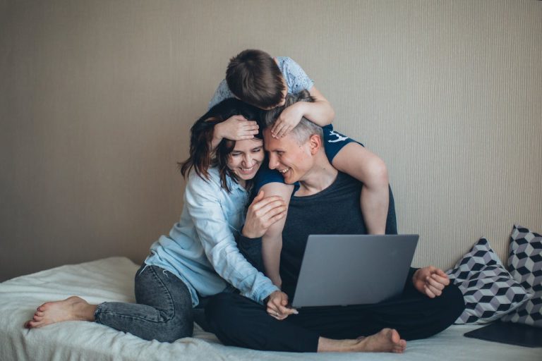Man working on a laptop at home while being affectionately embraced by his partner and child, illustrating balancing career and relationship.