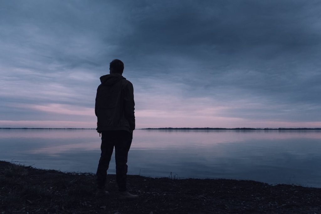 Man standing alone by a calm body of water at dusk, reflecting on choices between people-pleasing and self-respect.
