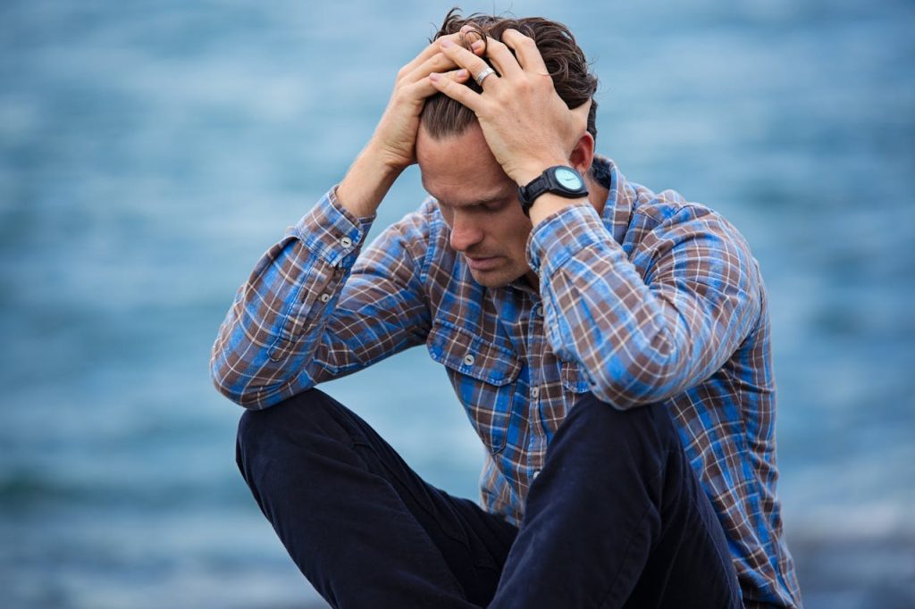 A young man sitting outdoors by the water, holding his head in frustration, illustrating internal emotional struggle and unmanaged anger in men.