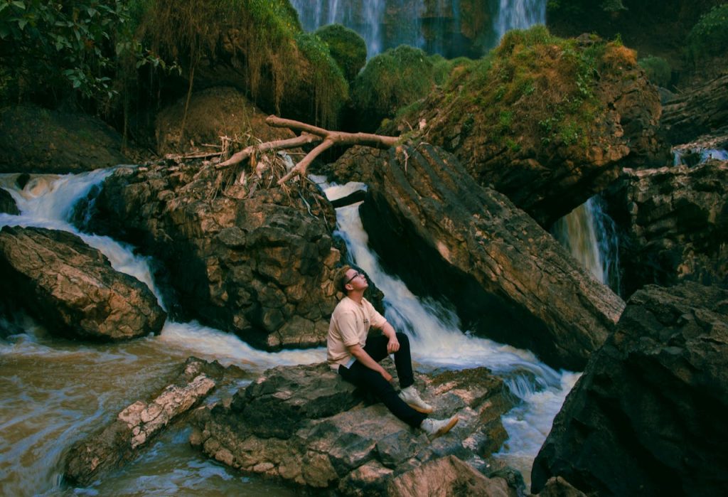 Man sitting calmly on rocks beside flowing water, immersed in stillness and reflection, symbolizing receptive feminine energy and inner awareness.