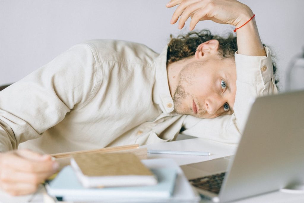 Man slumped at a desk with laptop and notebooks, appearing mentally exhausted from work overload and lack of personal boundaries.