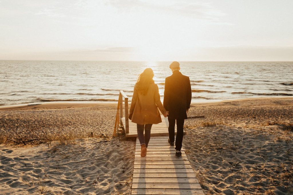 Couple walking hand in hand toward the sea at sunset, symbolizing emotional connection, shared direction, and balance between work and relationship.