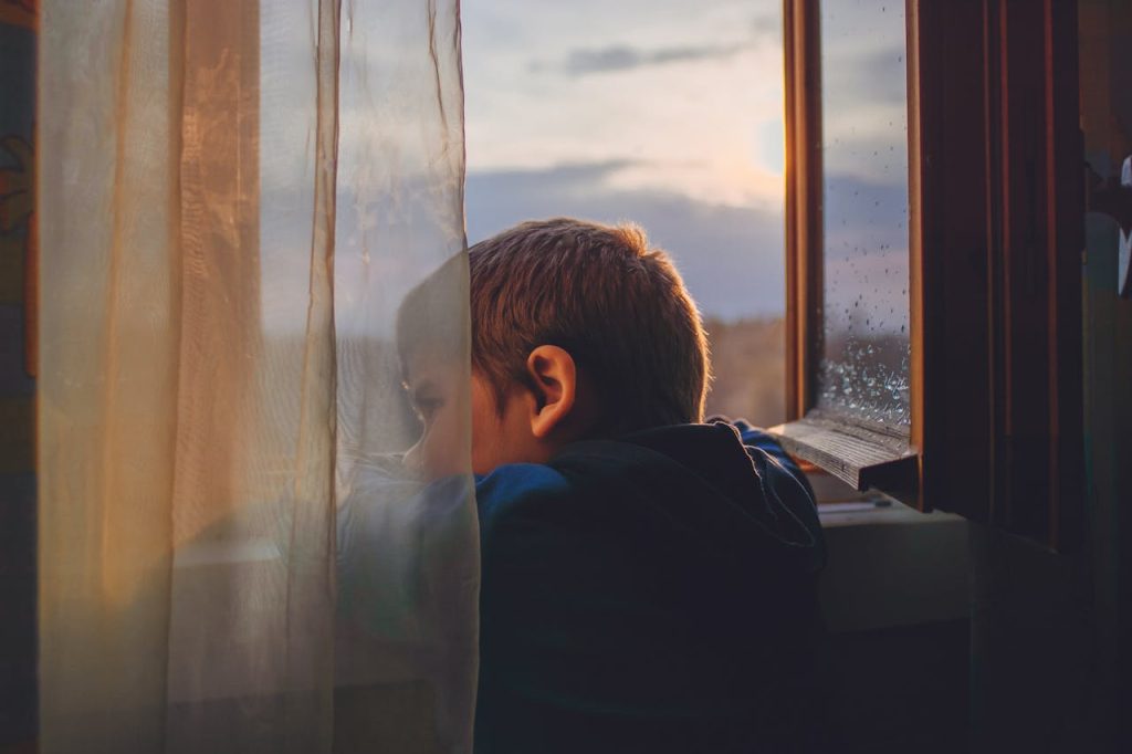 Young boy looking out a window alone, reflecting early experiences that shape the fear of being alone and comfort with solitude.