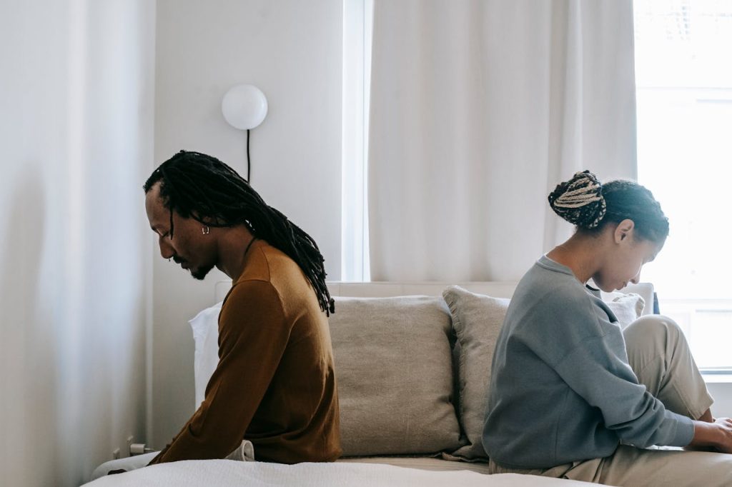 A couple sitting back-to-back on a bed, avoiding eye contact, illustrating emotional distance and intimacy difficulties often linked to unresolved childhood trauma in men.